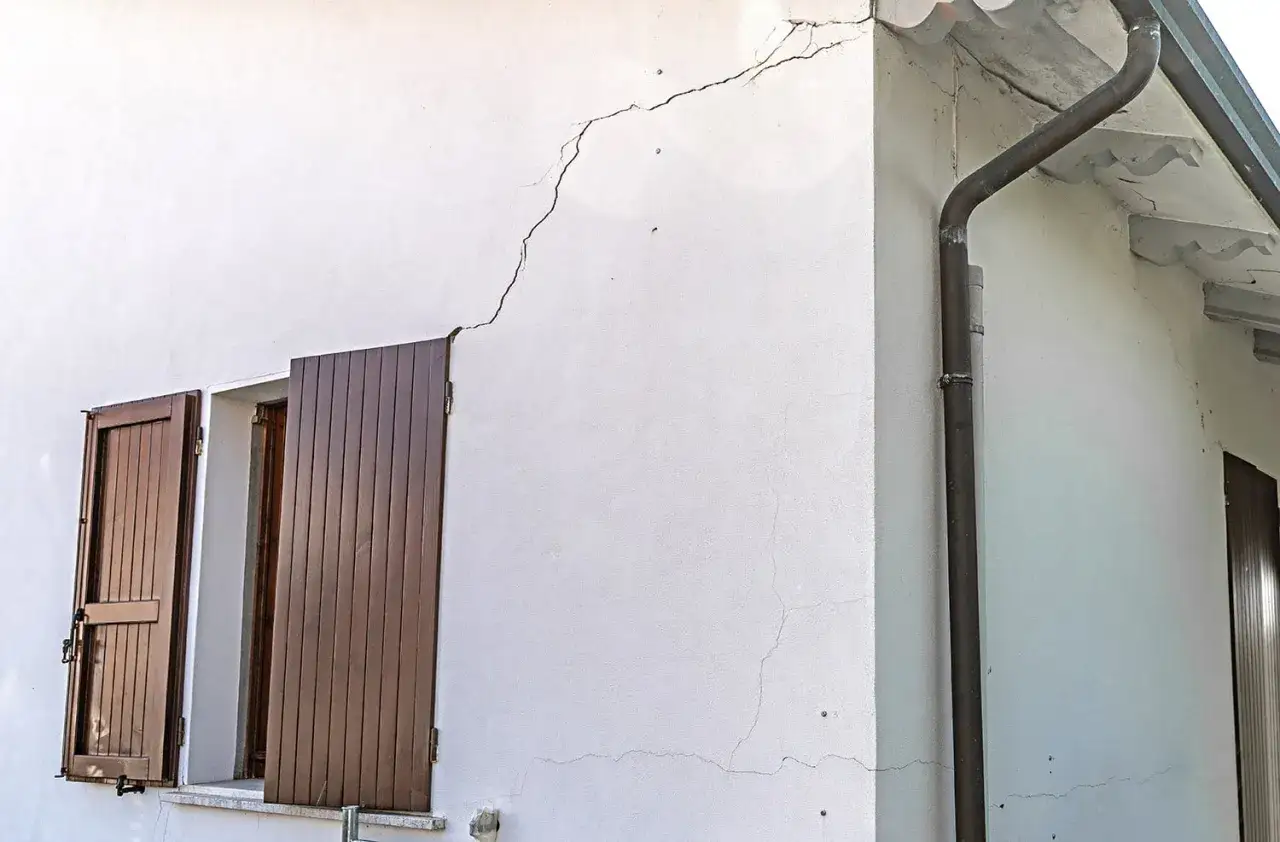 Dettaglio di un muro esterno bianco con ampie crepe visibili, che illustra come riempire le crepe nei muri esterni. Si vedono finestre con persiane marroni e una grondaia.