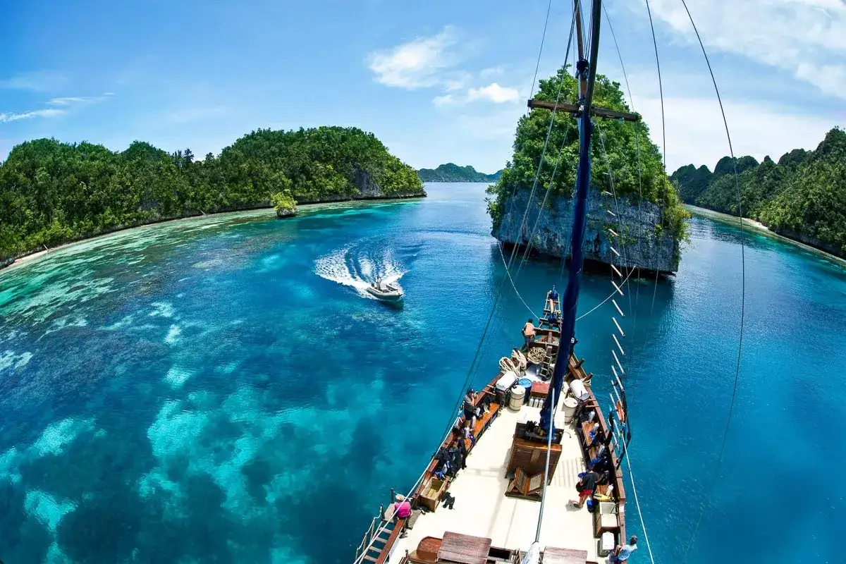 Segelboot in Raja Ampat, Indonesien. Die Inseln sind üppig grün, das Wasser ist kristallklar und türkisblau. Ein kleineres Boot fährt voraus.