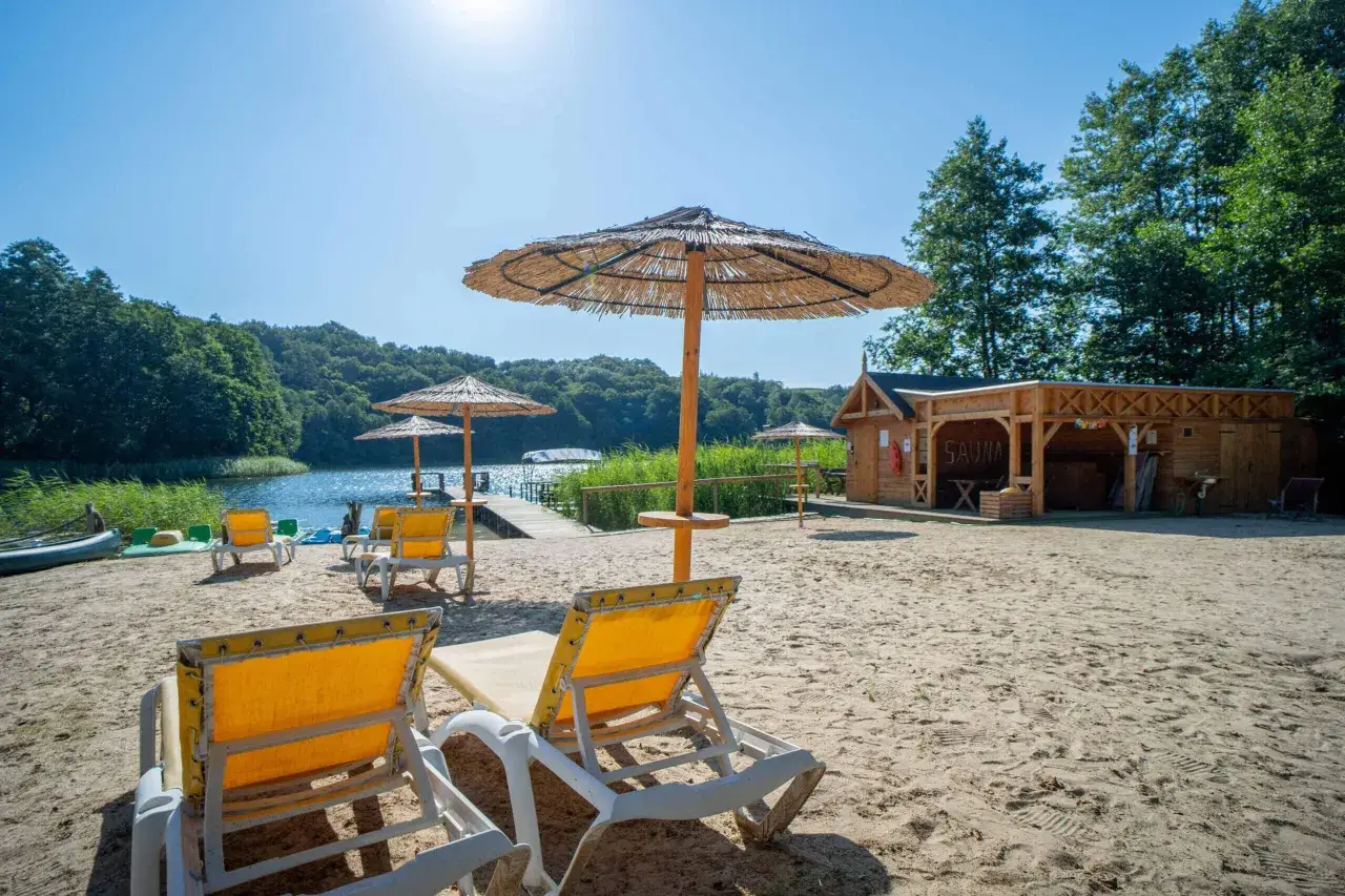 Plaża nad jeziorem z leżakami i parasolami. W tle drewniana sauna i pomost. Idealne miejsce na relaks nad wodą.