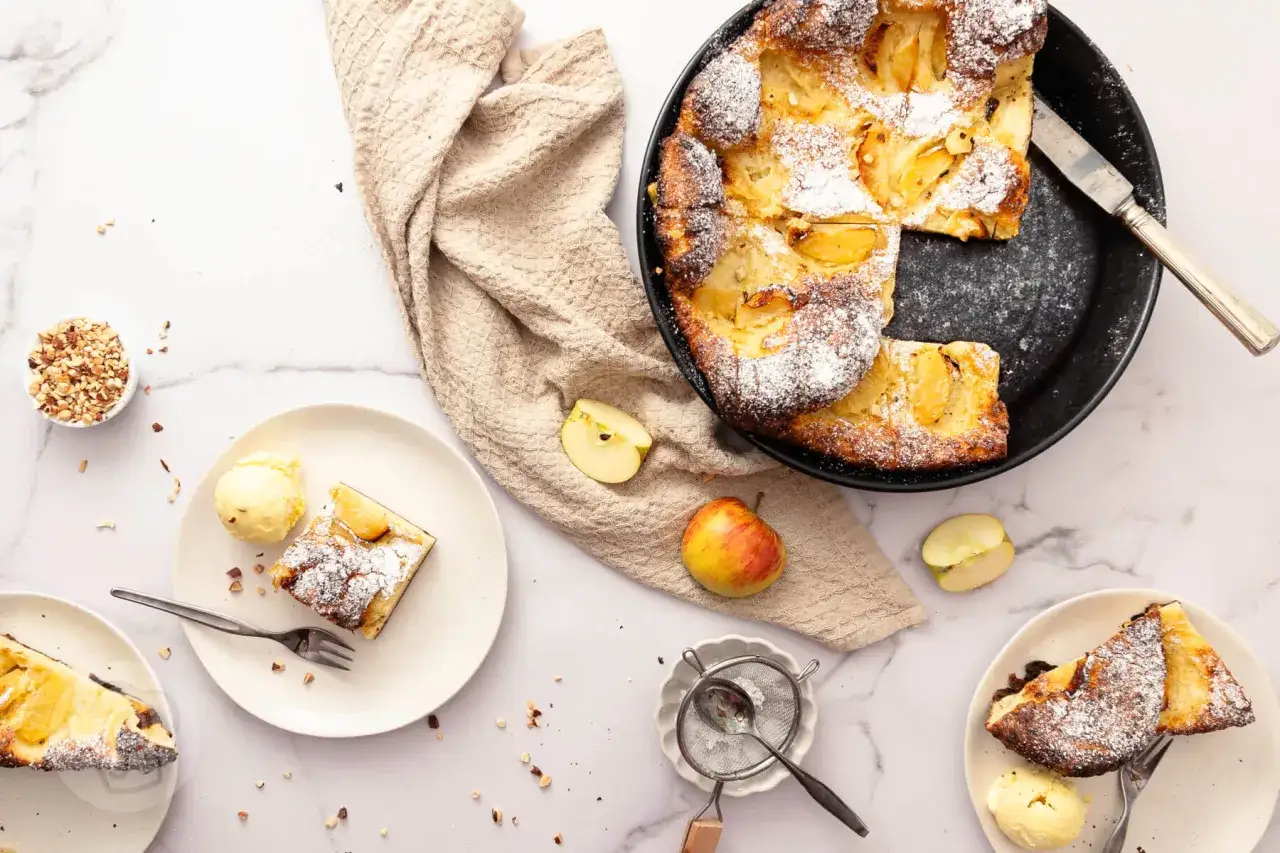 Saftige **Pfannkuchen vom Blech** mit Äpfeln, bestäubt mit Puderzucker, serviert mit Vanilleeis und gehackten Nüssen.