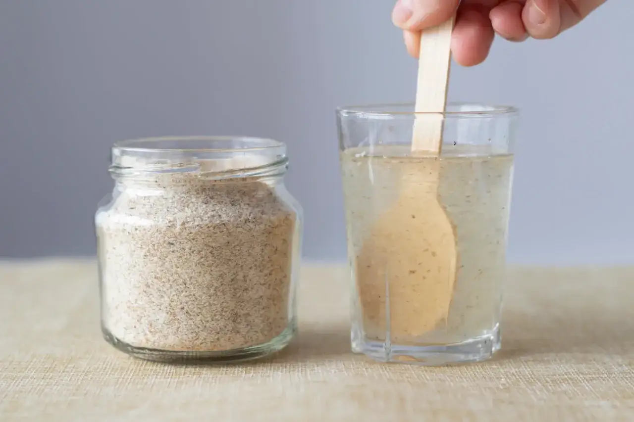 Glas mit Wasser und Holzstäbchen, daneben ein Glas mit Flohsamenschalen. Die richtige Flohsamenschalen Dosierung ist wichtig.