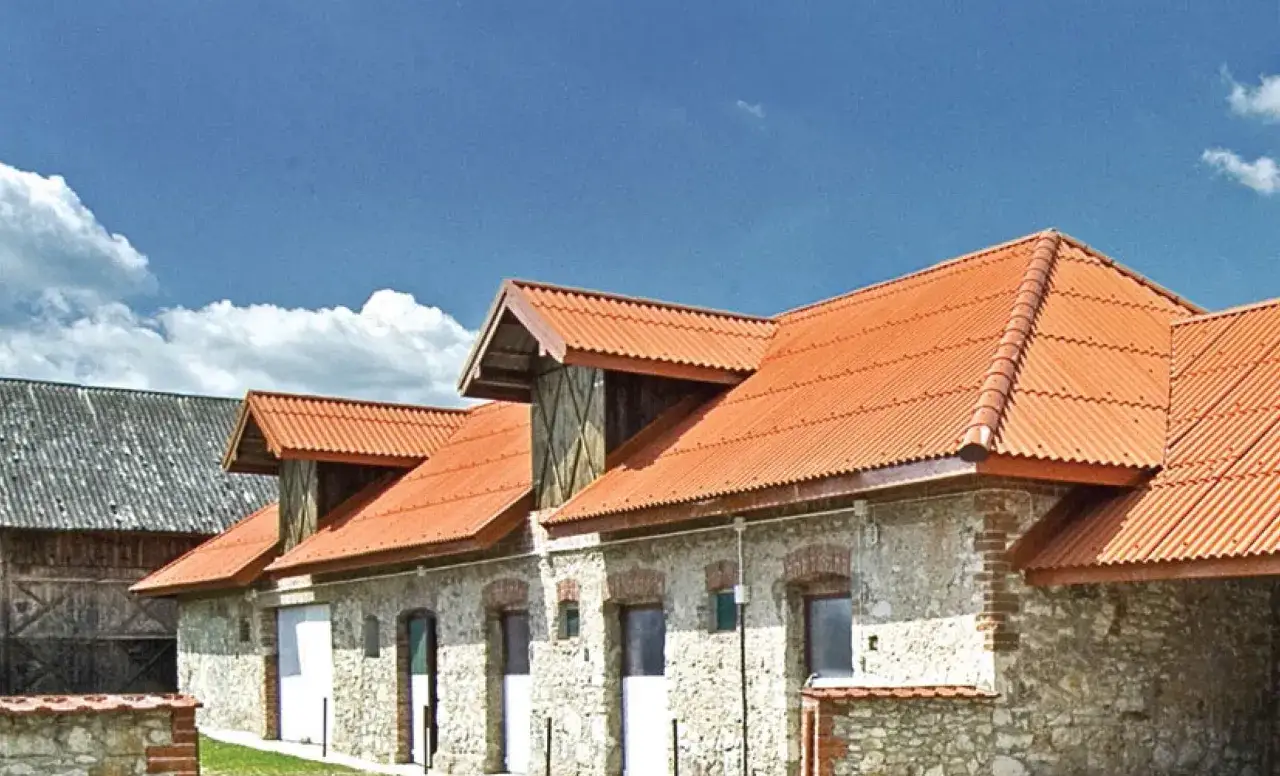 Stary budynek z szarym dachem, który może przypominać jak wygląda azbest, obok nowszego z pomarańczową, falistą blachą.