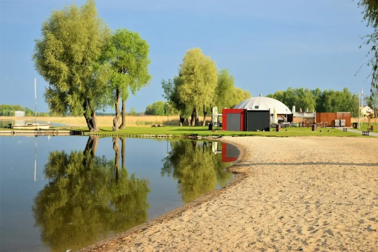 Zbąszyń atrakcje: piaszczysta plaża nad jeziorem, zacumowana łódka, zielone drzewa i nowoczesny budynek z kopułą.