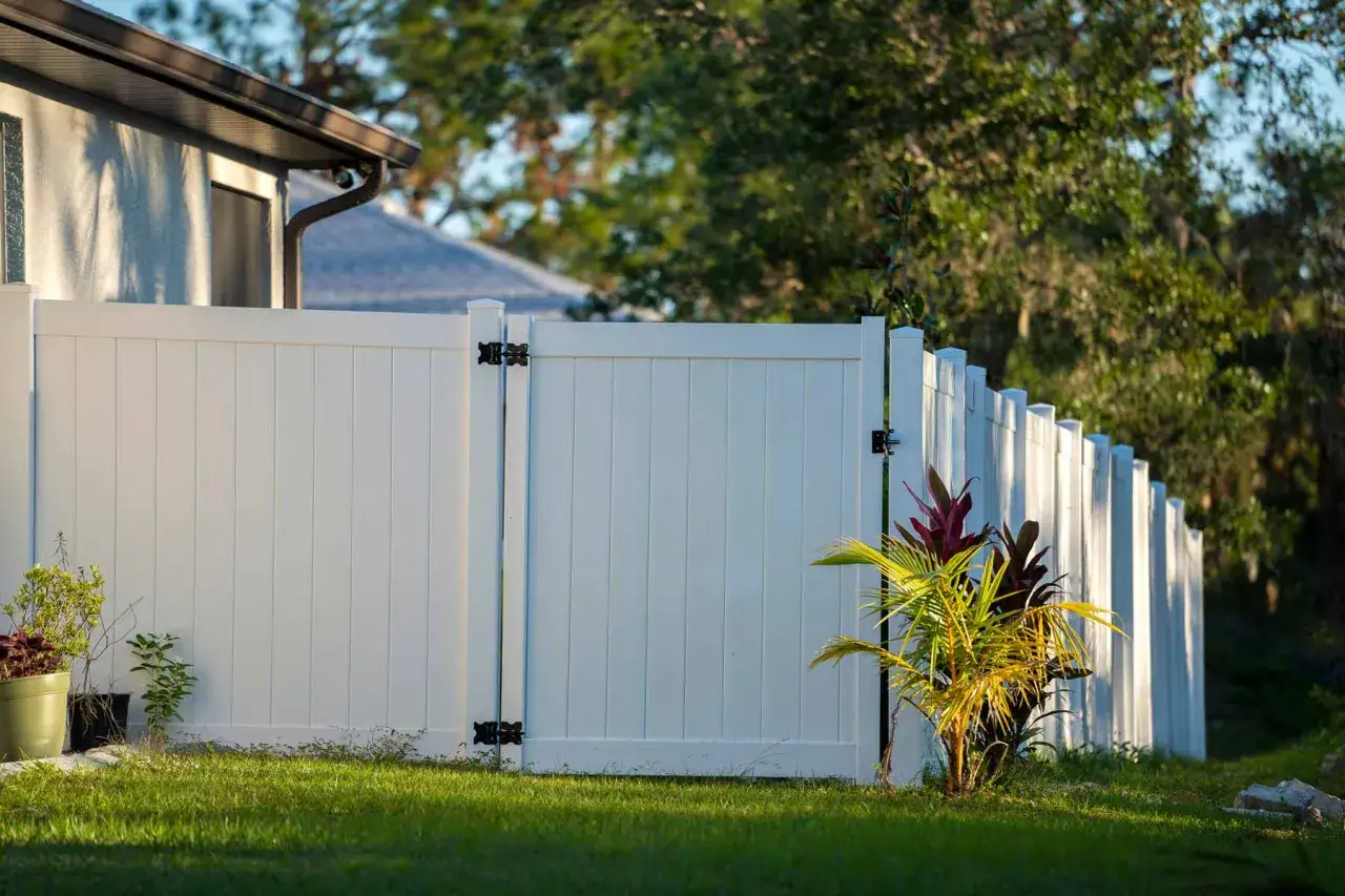 A newly completed vinyl fence installation surrounds a backyard, featuring a double gate and a single gate, with lush green grass and tropical plants.