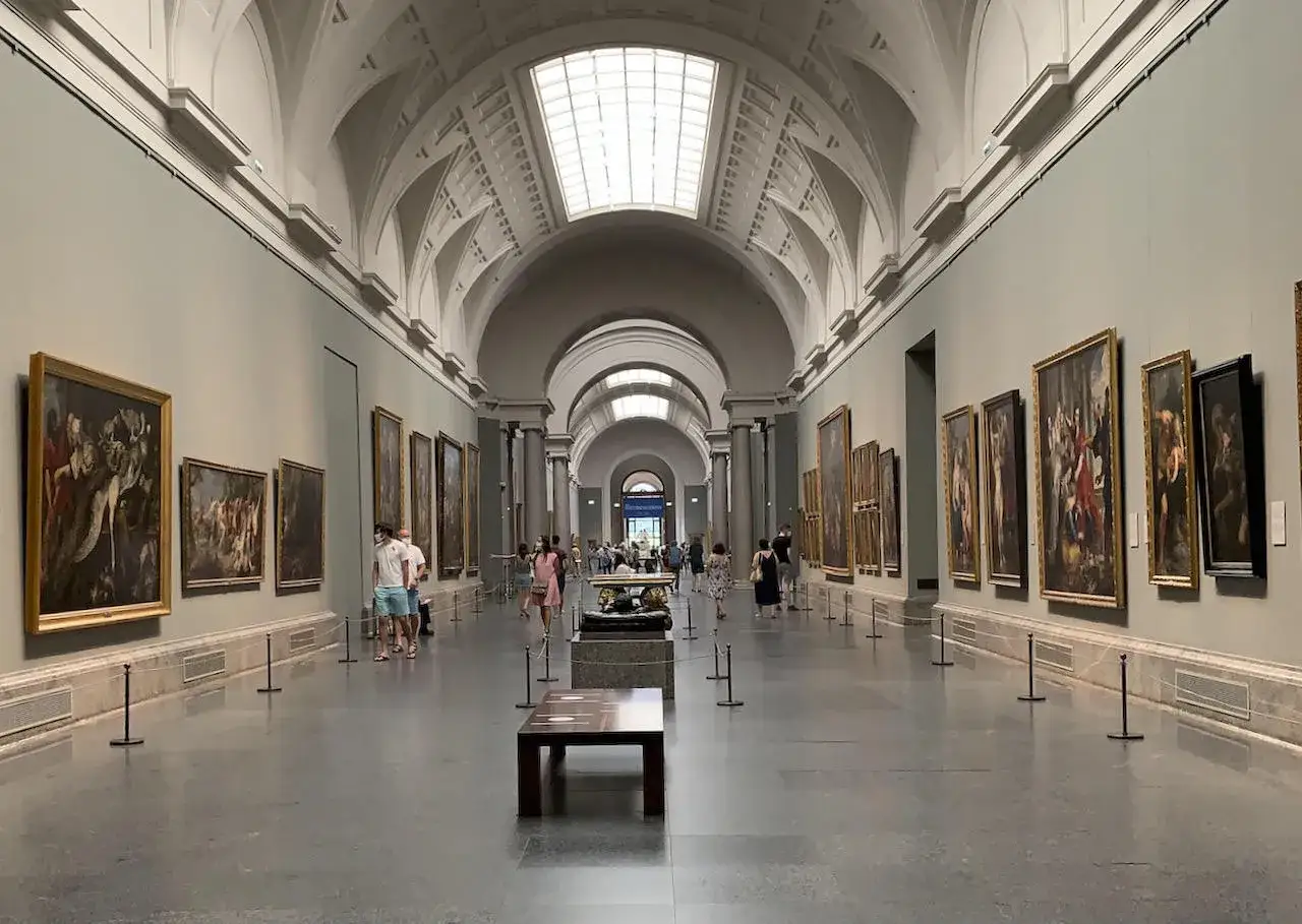 Długa, jasna sala pełna obrazów w złotych ramach. Ludzie spacerują po muzeum w Madrycie, podziwiając dzieła sztuki.