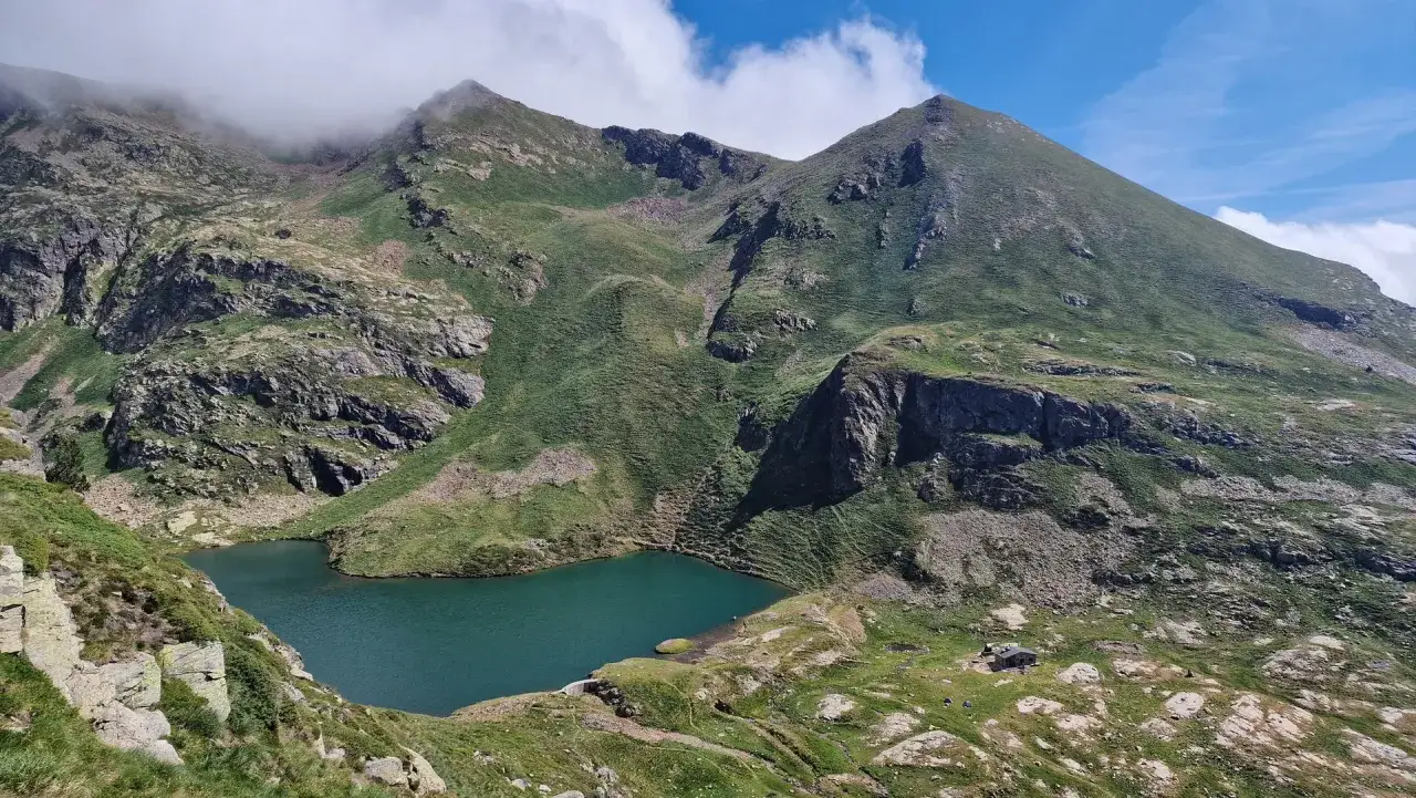Un lago turquesa en los Pirineos, rodeado de montañas verdes y rocosas. Un refugio solitario se asoma en la ladera, cerca de los coronallacs Andorra.