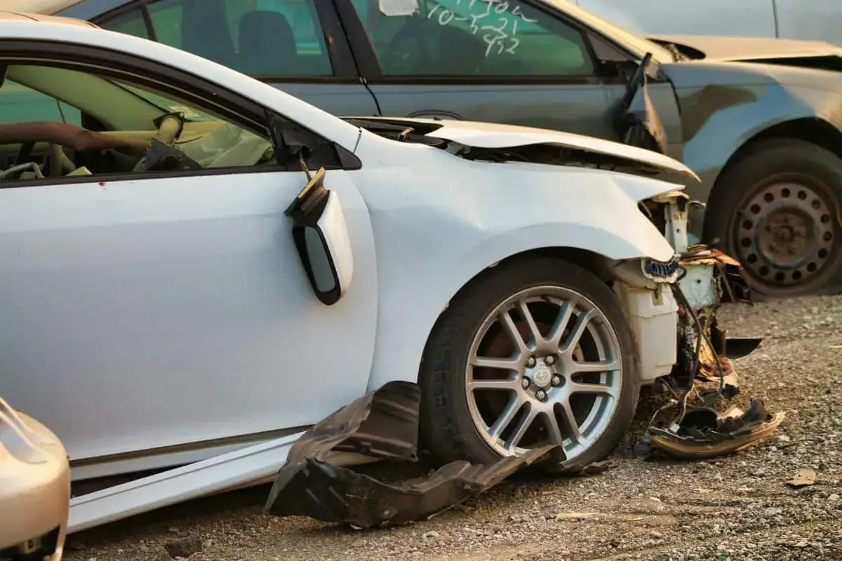 Uszkodzone białe auto na złomowisku. Zderzak i lusterko są wyrwane. Widać, jak działa ochrona zniżek po wypadku.