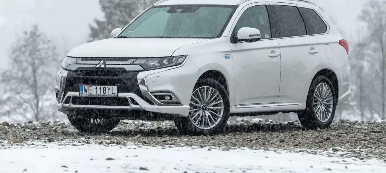 Biały Mitsubishi Outlander hybryda na kamienistym podłożu, w tle drzewa i padający śnieg.