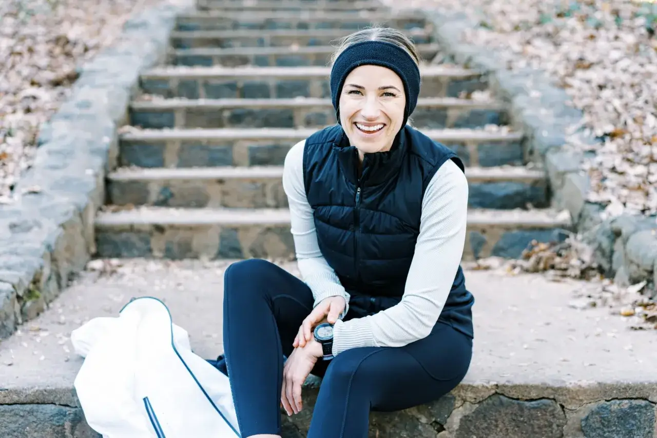 Lächelnde Frau in Laufkleidung, perfekt zum Joggen bei 10 Grad. Sie trägt eine Weste über einem Langarmshirt und eine Stirnband.