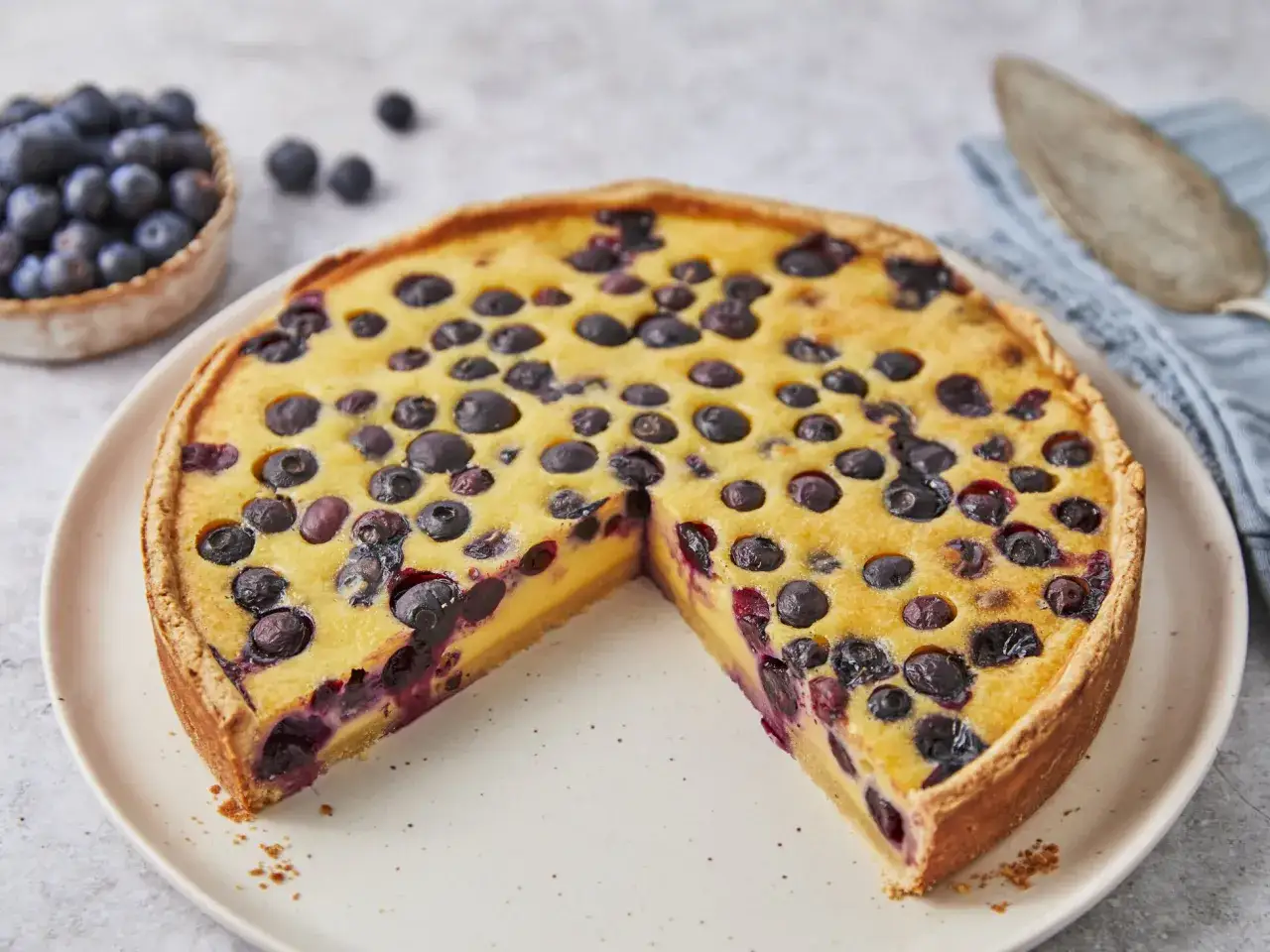 Ein köstlicher Heidelbeerkuchen mit Vanillepudding, belegt mit frischen Blaubeeren. Daneben eine Schale voller Blaubeeren.
