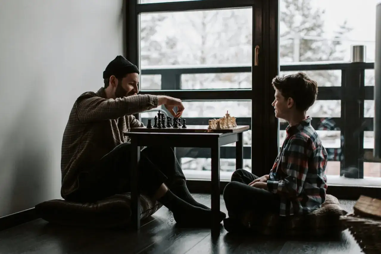 Ein Mann und ein Junge spielen Schach, ein Beispiel für gute Brettspiele. Sie sitzen auf dem Boden vor einem großen Fenster mit Blick auf eine verschneite Landschaft.