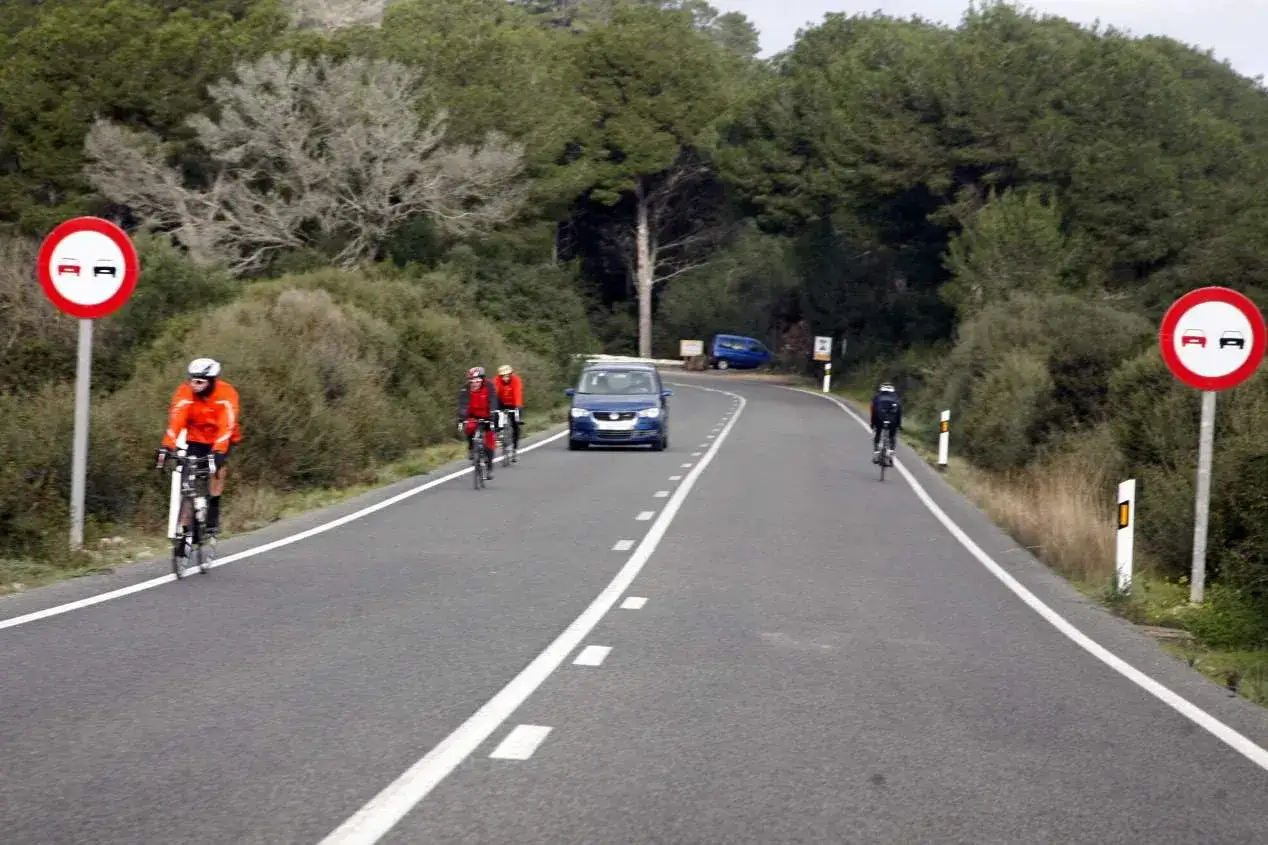 Ciclistas en carretera con señal de prohibido adelantar. ¿Es correcto este adelantamiento? Un coche azul circula en sentido contrario.