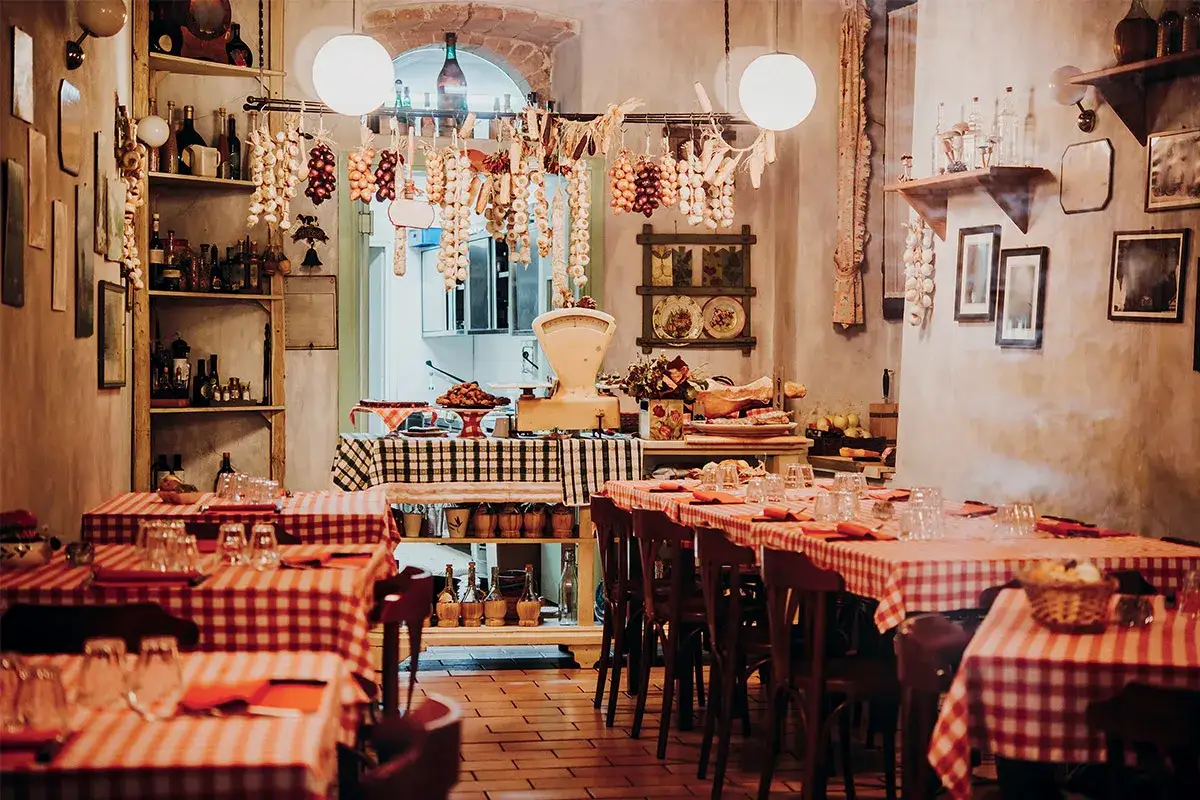 Interno di un'osteria o trattoria con tavoli apparecchiati, salumi appesi e un'atmosfera rustica.