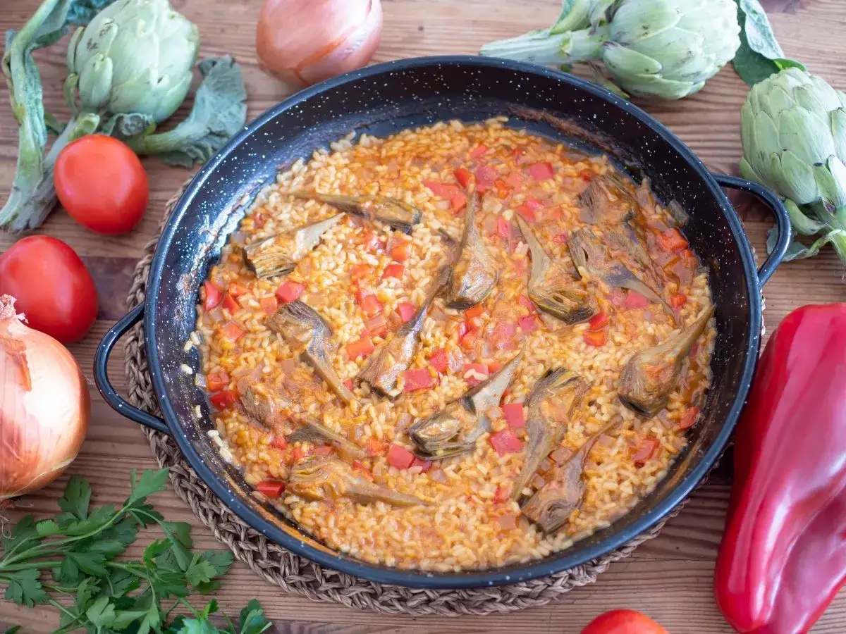 Arroz con alcachofas en paellera, rodeado de tomates, cebolla, pimientos y perejil. Un plato casero y delicioso.