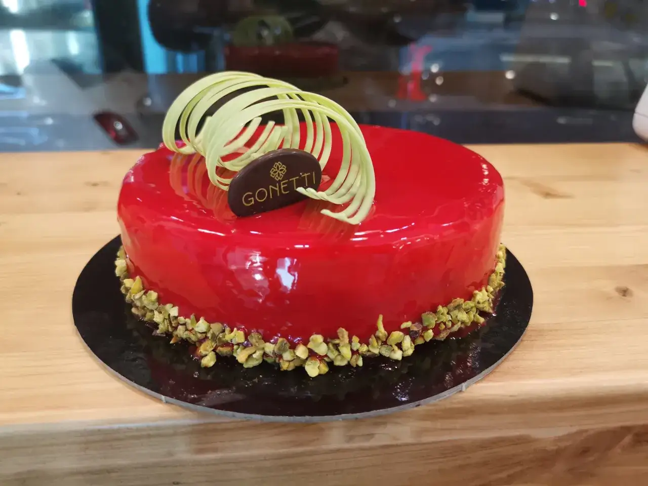 Torta rossa lucida con glassa a specchio lamponi, decorata con riccioli di cioccolato bianco e pistacchi tritati.