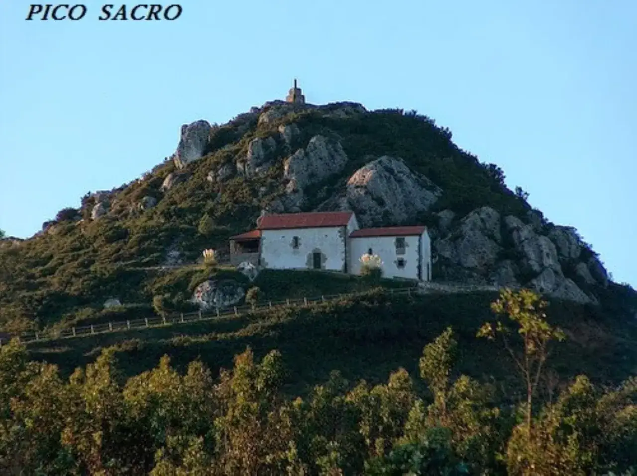Edificio blanco con tejado rojo en la ladera rocosa del Pico Sacro, bajo un cielo azul claro.
