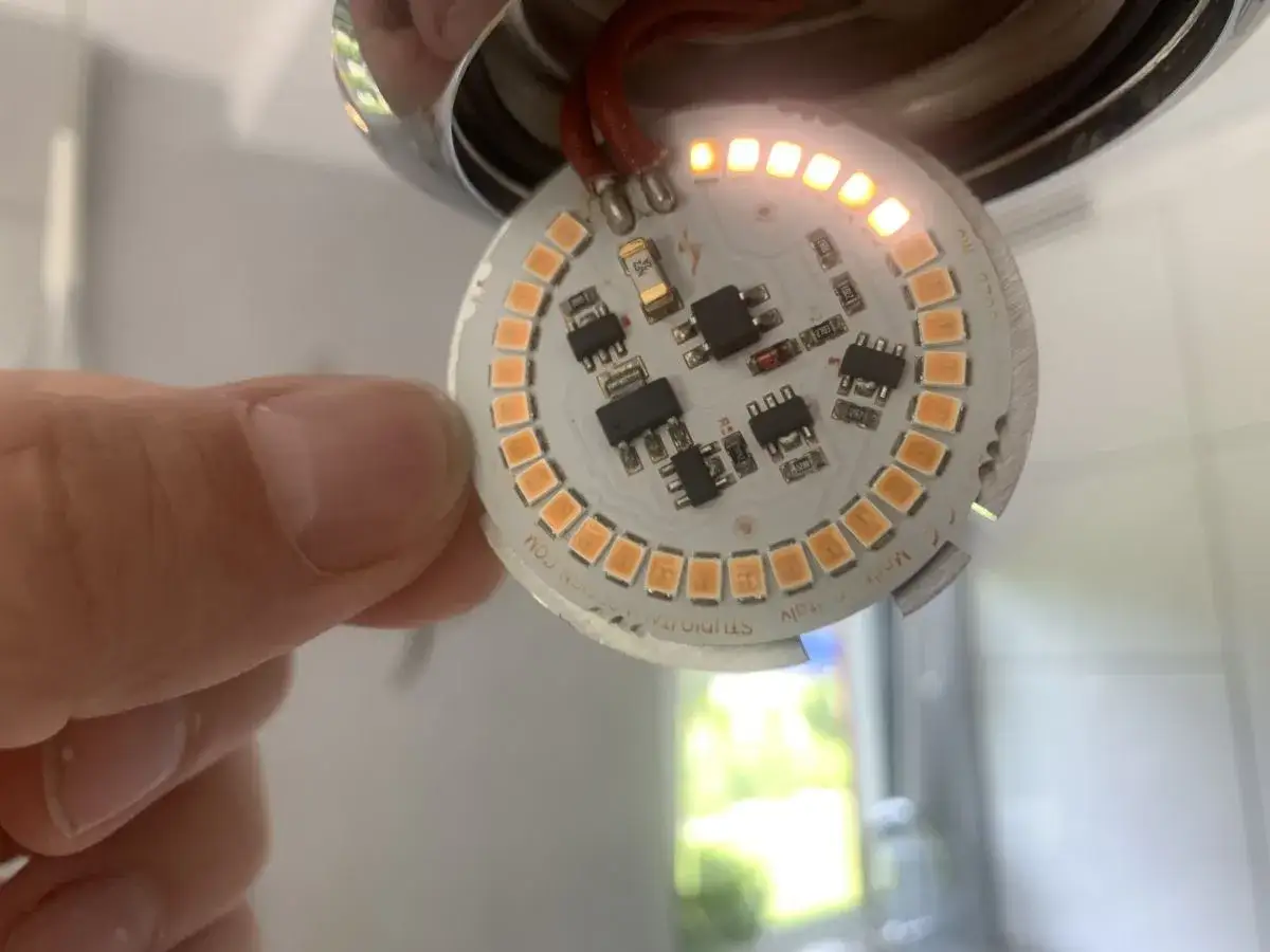 Naprawa lamp LED. Dłoń trzyma okrągłą płytkę z pomarańczowymi diodami i elementami elektronicznymi, część diod świeci.