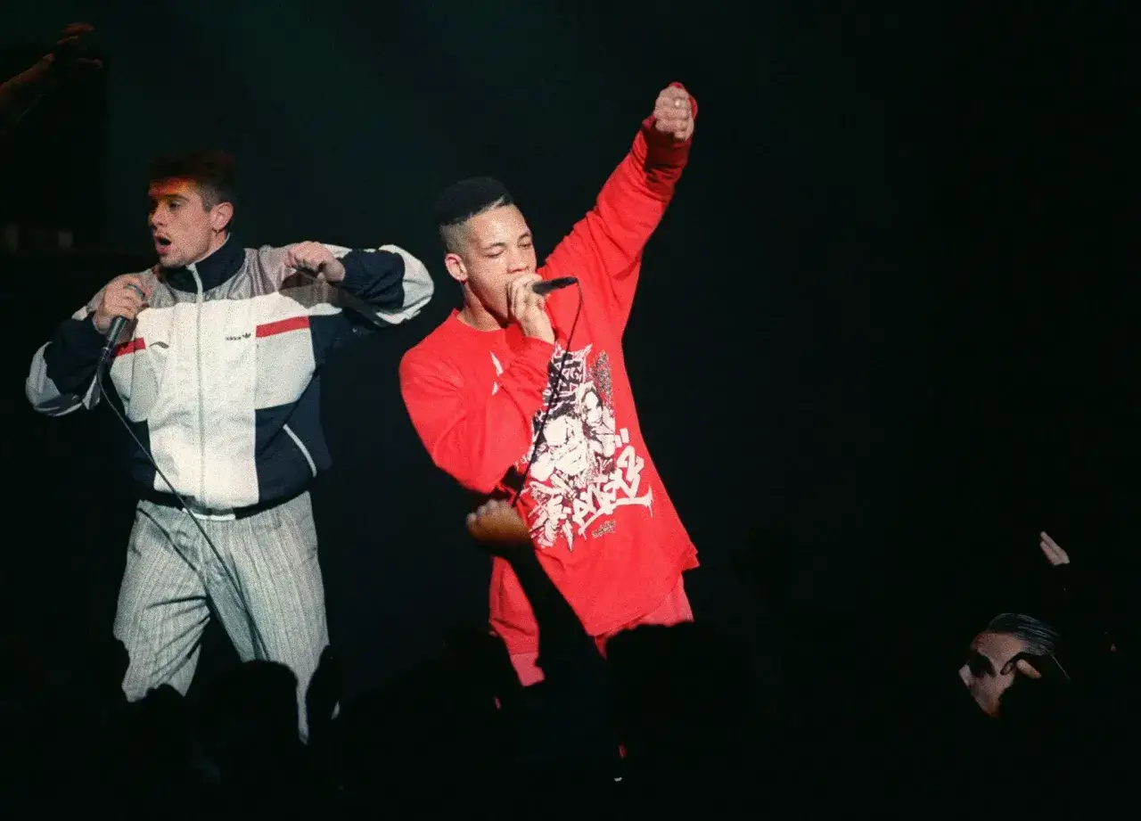 Deux rappeurs sur scène, l'un en rouge, l'autre en veste Adidas. Ils incarnent le **premier groupe de rap français** en pleine performance.