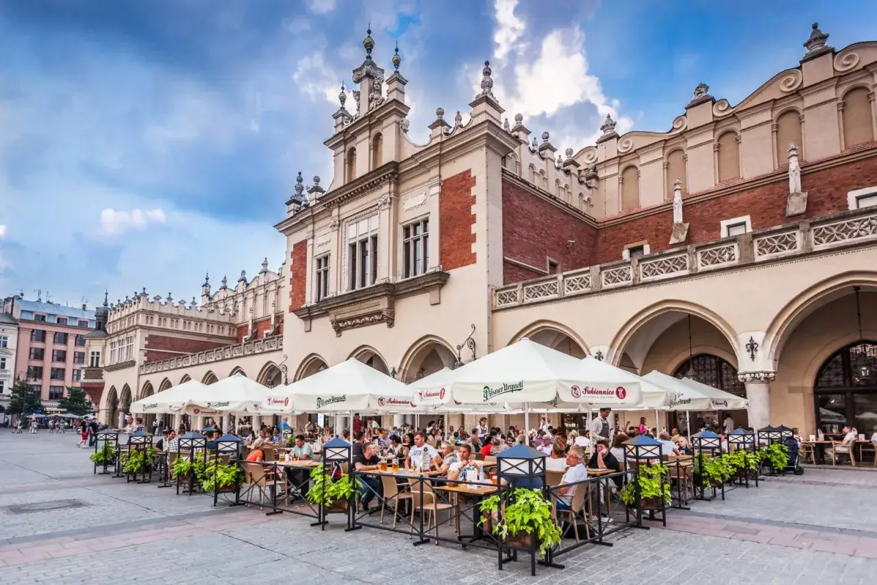 Tętniący życiem Rynek Główny w Krakowie, największy rynek w Polsce, z ogródkami restauracyjnymi i Sukiennicami w tle.