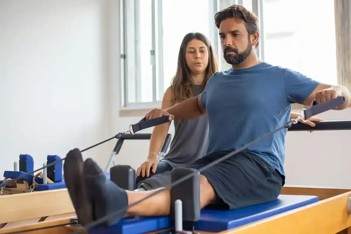 Mann trainiert auf Pilates-Gerät mit Trainerin. Frage: Was bezahlt die Krankenkasse?