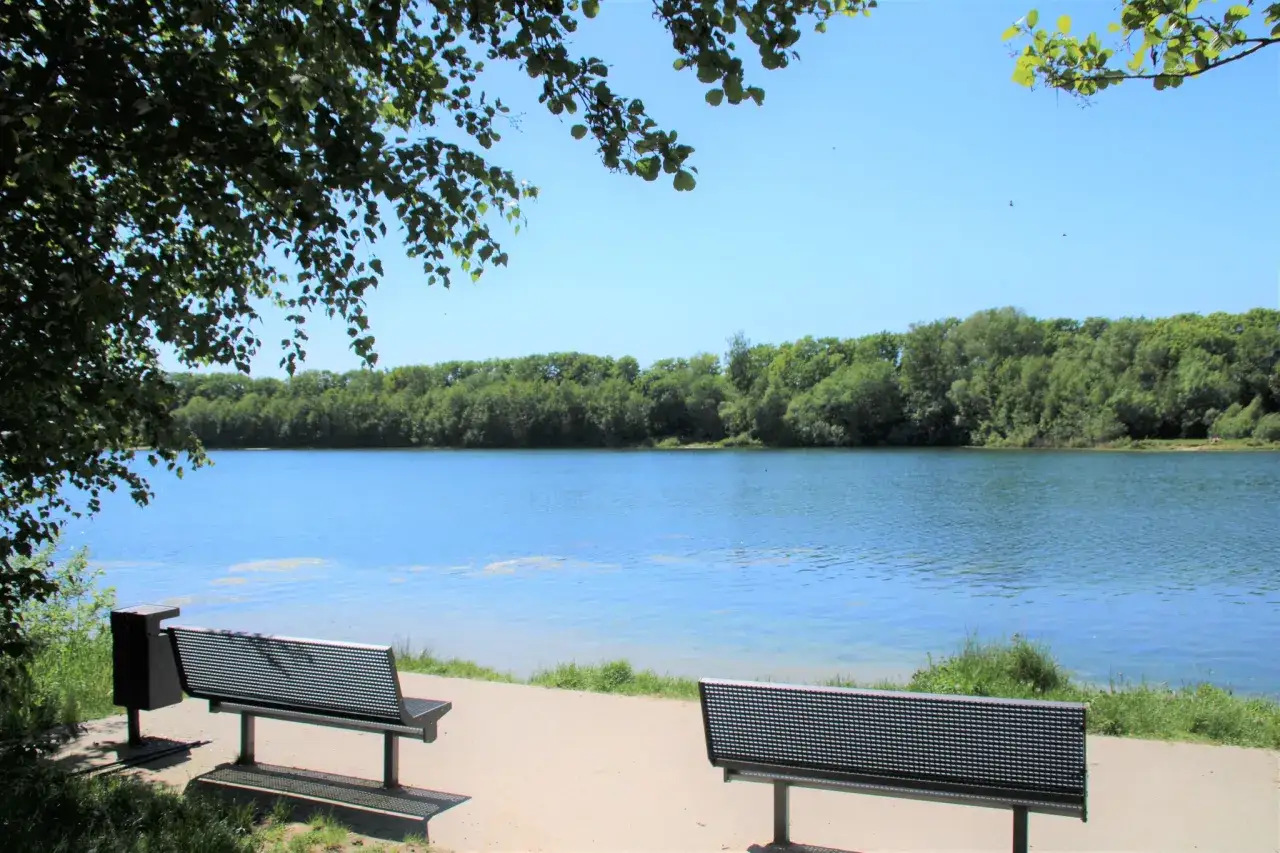 Zwei B&auml;nke laden zum Verweilen am Ufer des halener Badesees ein. B&auml;ume s&auml;umen das klare Wasser unter blauem Himmel.