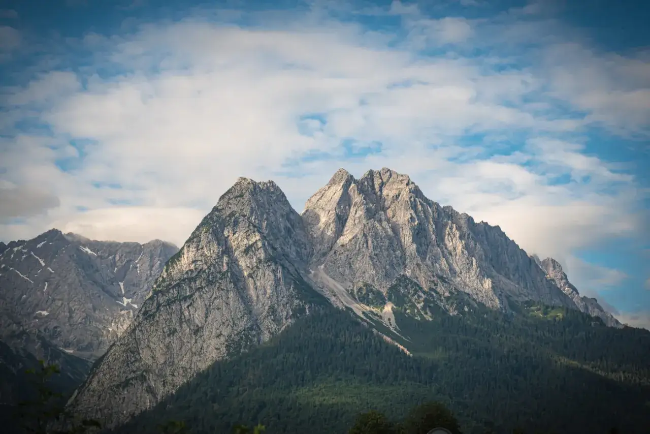 Majestatyczne szczyty Zugspitze, najwyższego szczytu Niemiec, wznoszą się ponad gęstym lasem, a błękitne niebo zdobią białe chmury.