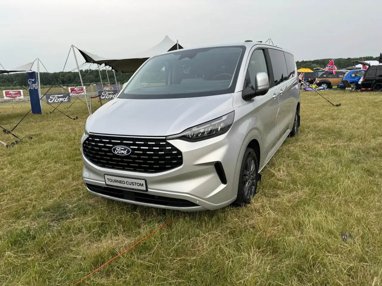 Srebrny Ford Tourneo Custom, jaki bus z napędem 4x4, stoi na trawie obok namiotów Forda.