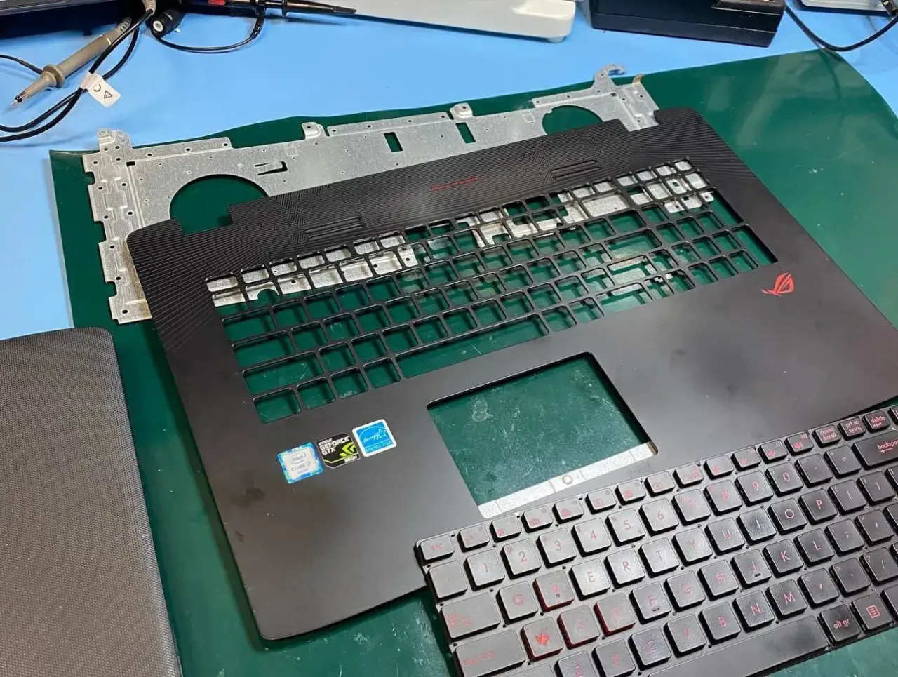 Rozpoczęta wymiana klawiatury w laptopie. Widoczna górna obudowa z pustymi miejscami na klawisze i nowa klawiatura z czerwonymi oznaczeniami.