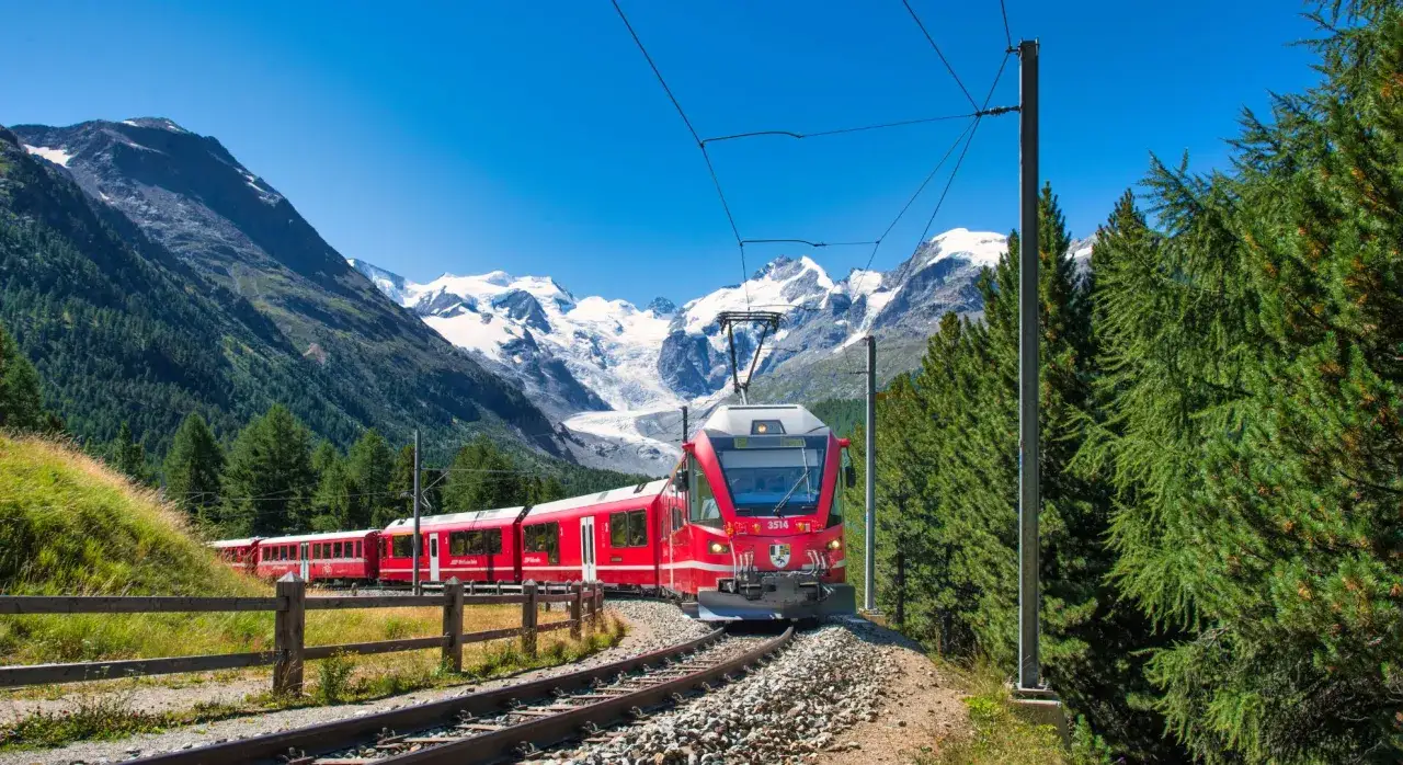 Czerwony pociąg pnie się po torach w Alpach. W tle majestatyczne, ośnieżone szczyty i lodowiec. To niezapomniana podróż koleją w Szwajcarii.