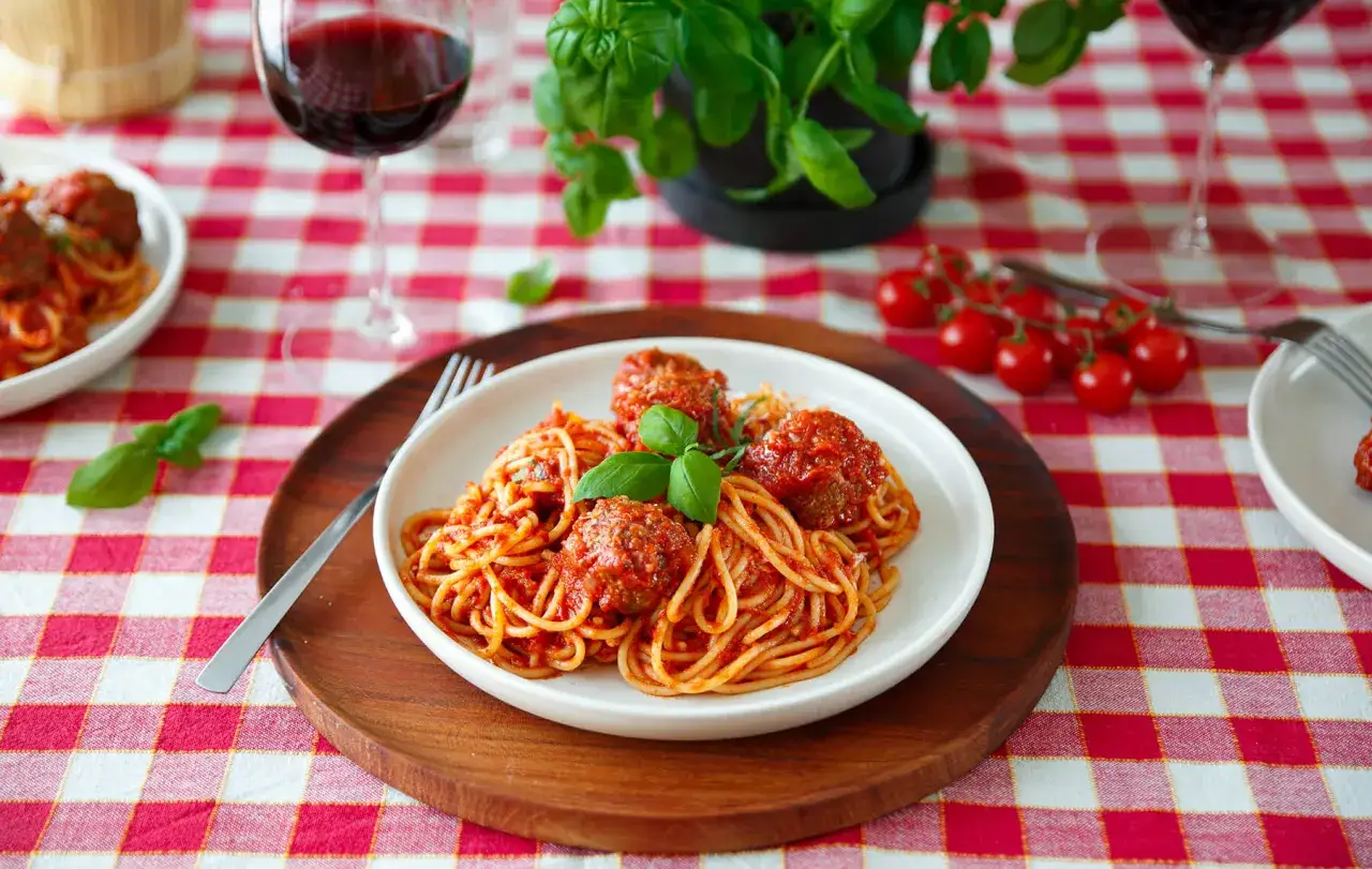 En tallrik med spaghetti och k&ouml;ttbullar (polpette) i tomats&aring;s, garnerad med basilika. R&ouml;d rutig duk och ett glas r&ouml;dvin i bakgrunden.