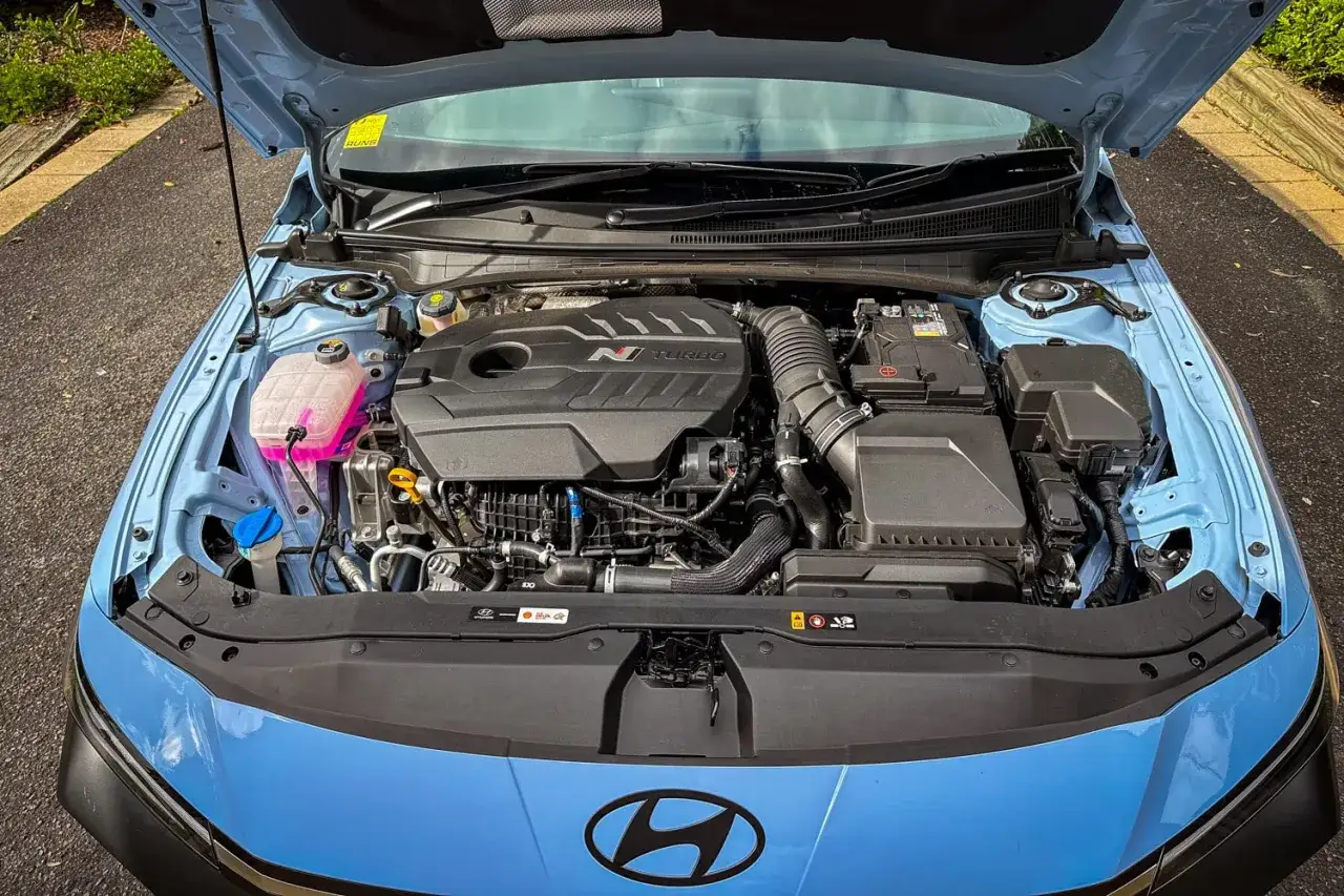 El motor i30n turbo, listo para la acci&oacute;n. Refrigerante rosa brillante y componentes negros pulcros bajo el cap&oacute; abierto de un coche azul.