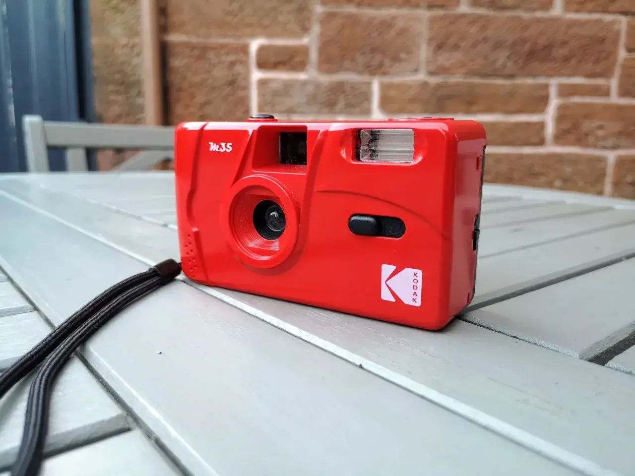 Czerwony aparat Kodak M35 z czarną smyczą leży na szarej, drewnianej ławce.