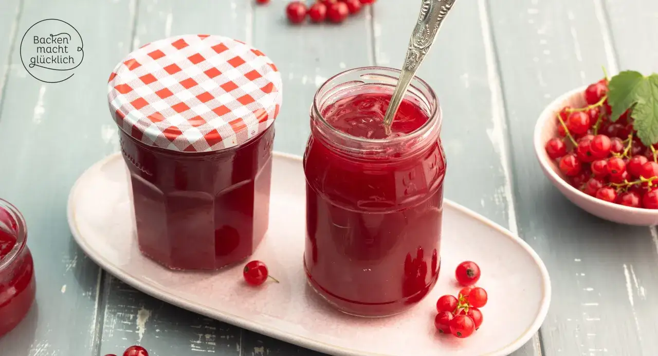 Zwei Gläser mit leuchtend roter, einfacher Johannisbeermarmelade, daneben frische Johannisbeeren und ein Löffel.