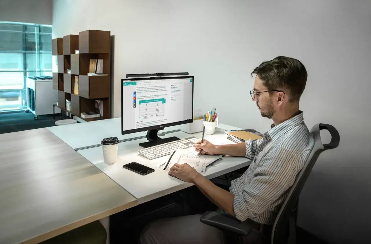 Un homme concentré travaille sur son ordinateur, prenant des notes sur papier. Une tasse de café à portée de main, il incarne une approche 