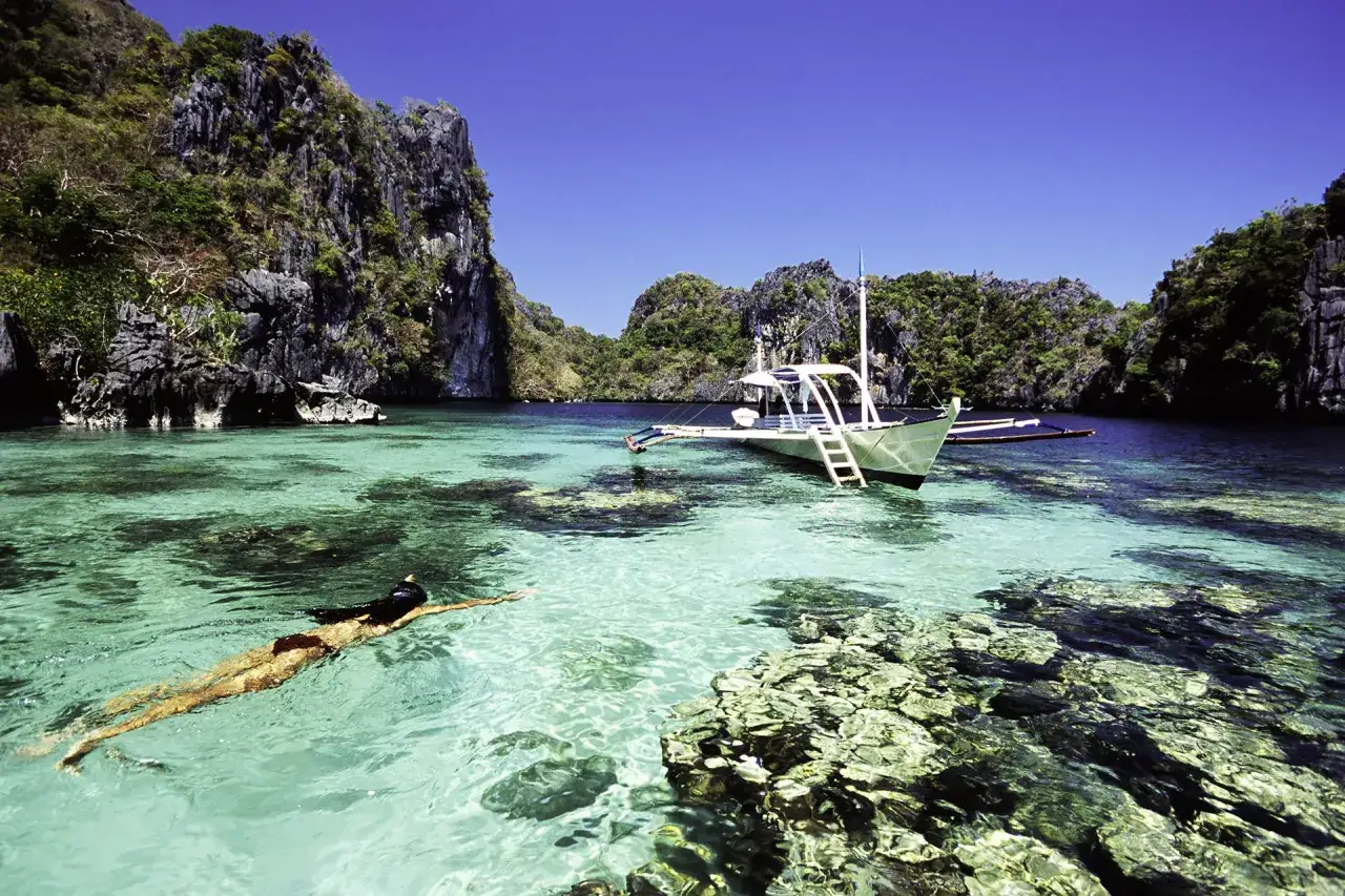 Una persona nada en aguas cristalinas, con un barco tradicional filipino anclado cerca. ¡La aventura de viajar a Filipinas te espera!