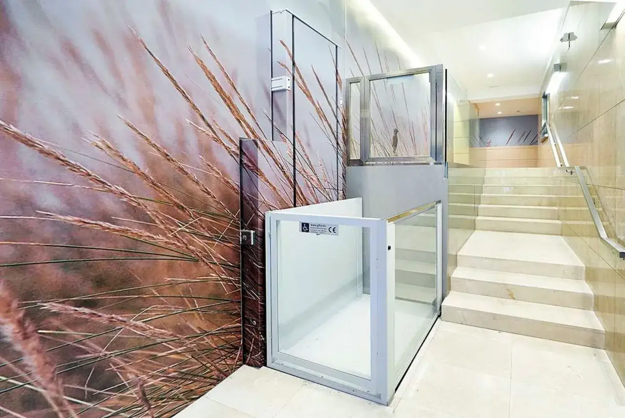 Un portal moderno y accesible. Una pared con un mural de hierbas secas inspira ideas sobre como decorar un portal de vecinos, junto a una plataforma elevadora de cristal.