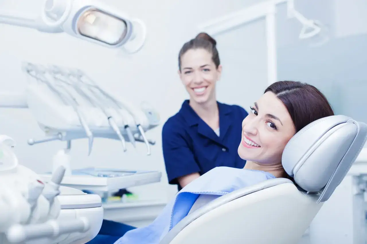 Uśmiechnięta pacjentka na fotelu dentystycznym, za nią uśmiechnięta dentystka.