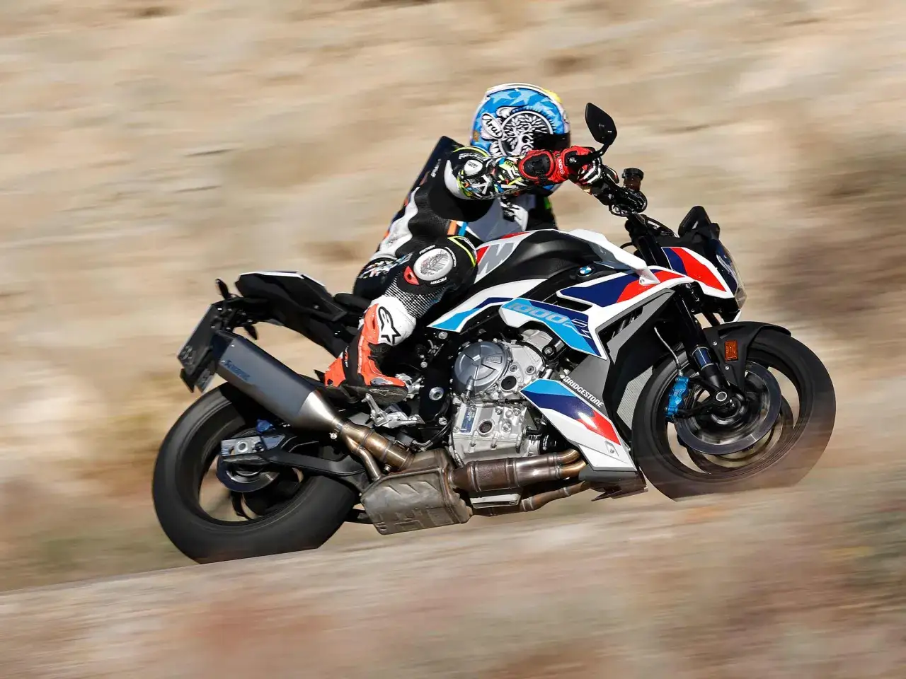 Motocyklista w kasku i kombinezonie pędzi na sportowym motocyklu BMW M1000R po piaszczystej drodze.