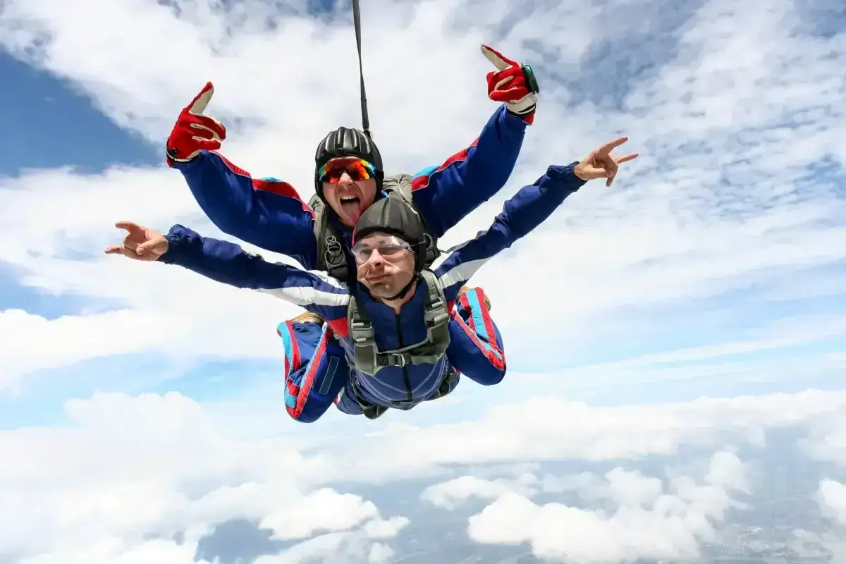 Twee parachutisten maken een tandem sprong. De voorste springt met een brede grijns, de achterste met een rock-'n-roll gebaar. Dit is wat HSS (High Speed Skydiving) is!
