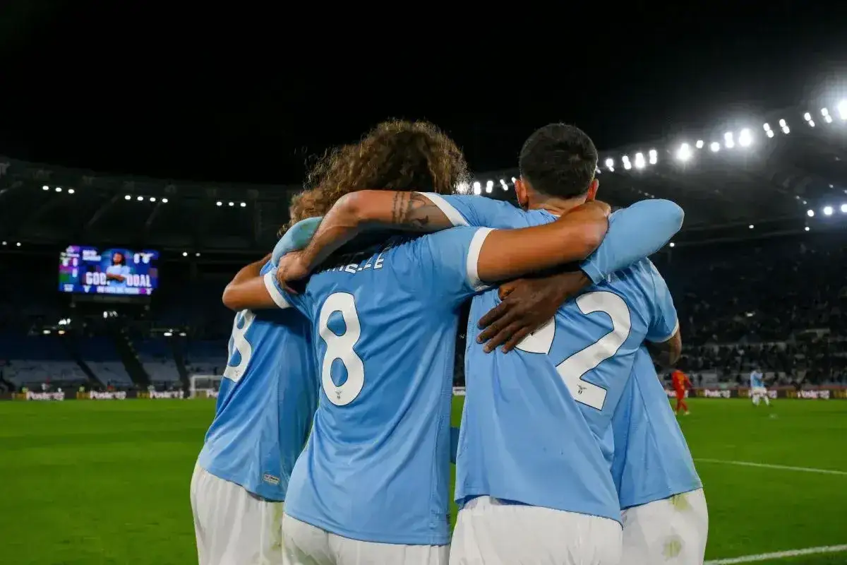 Piłkarze Lazio świętują bramkę w meczu z A.C. Milan. Stadion wypełniony kibicami, nocna atmosfera.