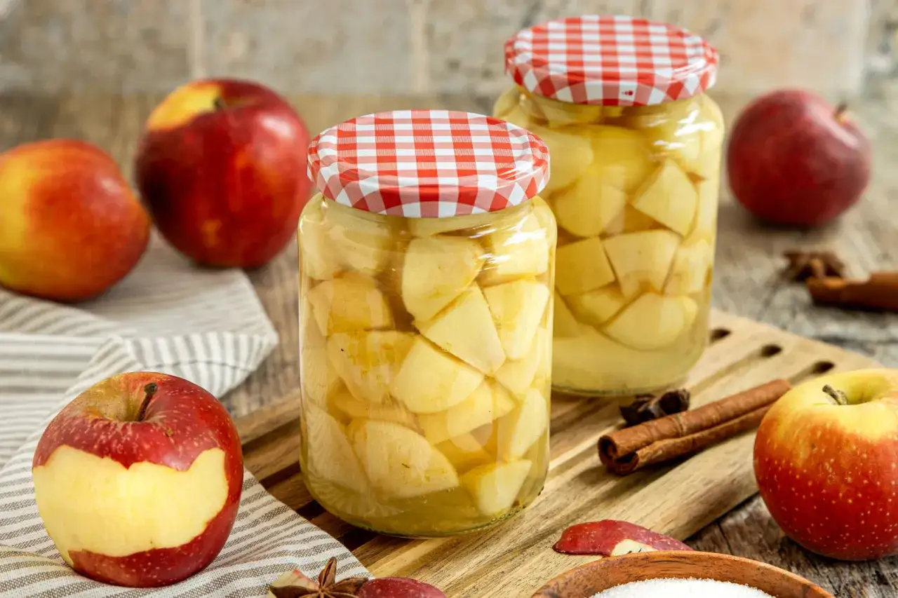 Zwei Gläser mit Apfelstücken, bereit zum Apfel einkochen. Frische Äpfel, Zimtstangen und Sternanis liegen daneben.