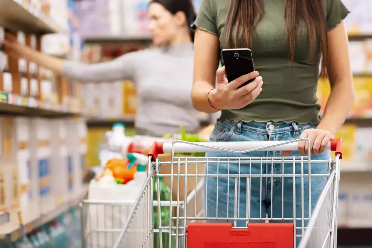 Kobieta z telefonem w ręku pcha wózek sklepowy, robiąc zakupy w supermarkecie. To typowy obrazek z branży retail, gdzie technologia ułatwia konsumentom codzienne czynności.