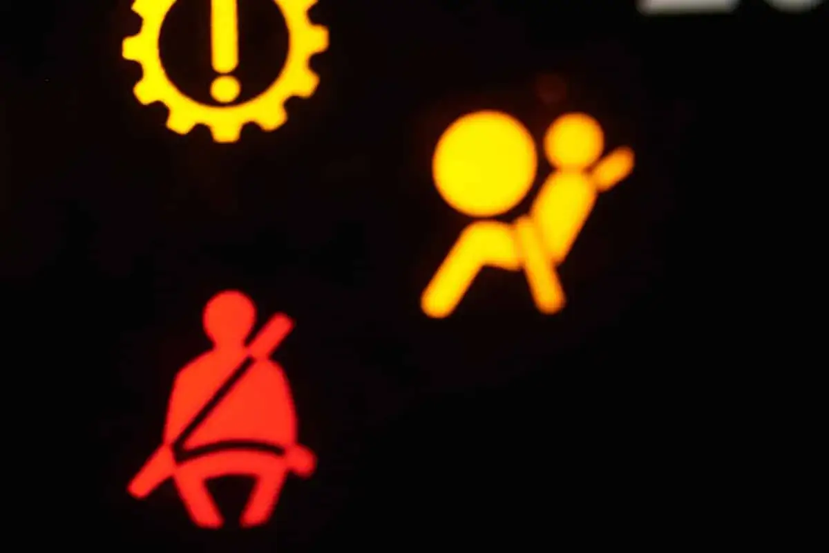 Das gelbe airbag symbol leuchtet, zusammen mit dem Symbol für den nicht angelegten Sicherheitsgurt. Ein gelbes Zahnrad mit Ausrufezeichen ist ebenfalls sichtbar.