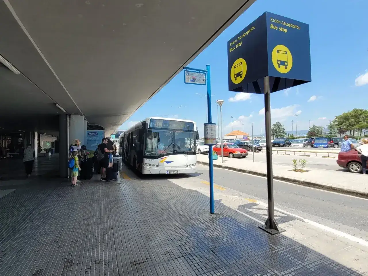 Autobus na Chalkidiki lotnisko czeka na pasażerów. Ludzie z bagażami stoją pod zadaszeniem.