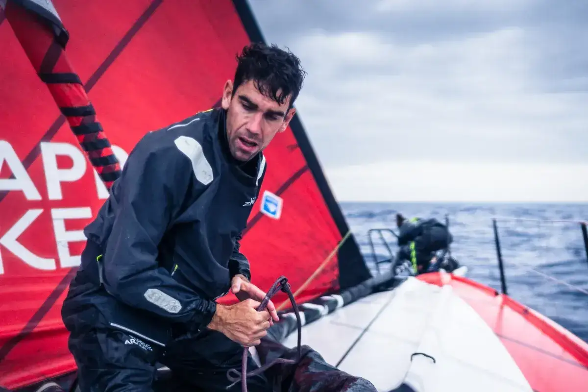 Gaston Morvan, concentr&eacute;, man&oelig;uvre une voile rouge sur un voilier en pleine mer. L'oc&eacute;an est agit&eacute; sous un ciel couvert.