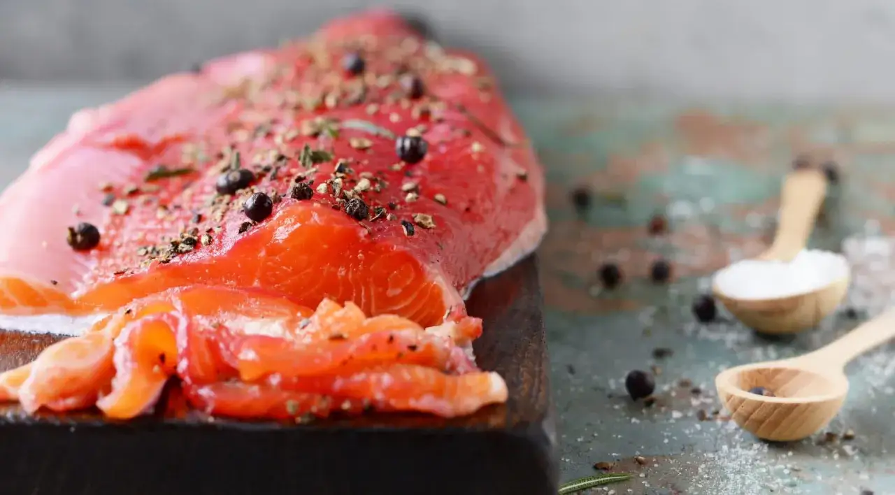Un filet de truite fraîchement préparé pour un gravlax, assaisonné de poivre et d'herbes. Deux cuillères en bois contiennent du sel et des baies de genièvre.