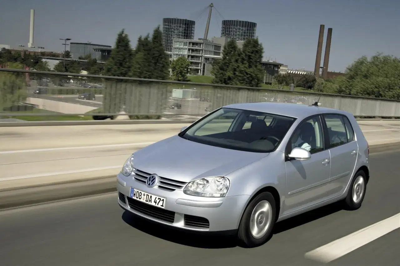 Srebrny Volkswagen Golf 5 z 2004 roku jedzie po drodze z nowoczesnymi budynkami w tle.