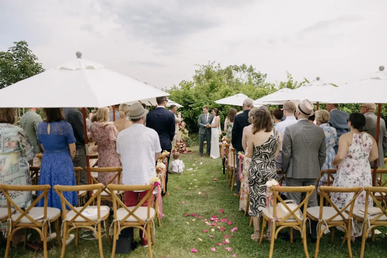Hochzeit im Weingut: Gäste sitzen auf Stühlen unter Sonnenschirmen, während das Brautpaar vor einer üppigen Kulisse steht.