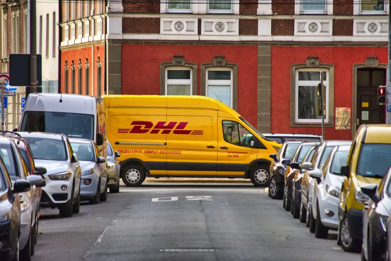 Żółta furgonetka DHL na ulicy, obok zaparkowane samochody. Zastanawiasz się, ile zarabia kurier?