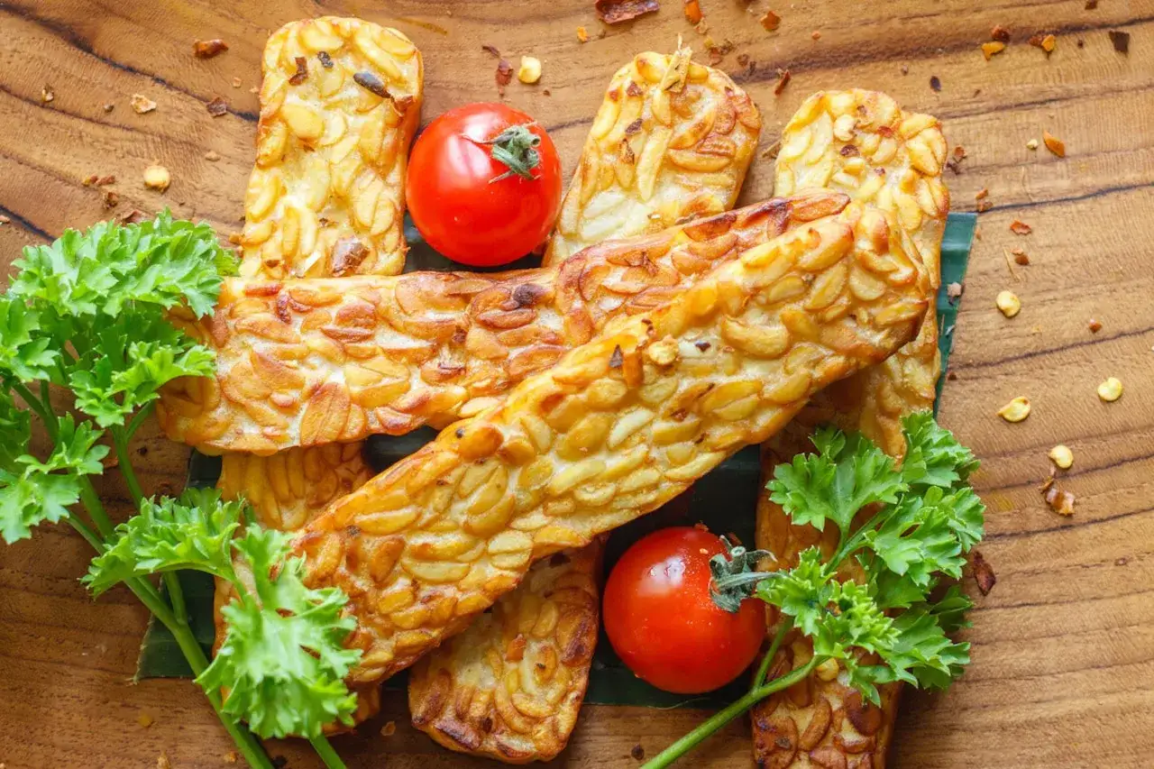 Usmażony tempeh, czyli co to jest? Złociste kawałki na drewnianej desce, ozdobione pomidorkami i natką pietruszki.